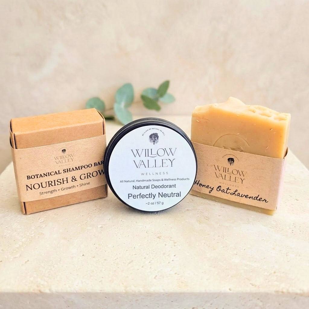 Three Willow Valley Wellness products arranged on a light stone surface: a botanical shampoo bar in a kraft box, a round tin of natural deodorant labeled perfectly neutral, and a honey oat lavender soap bar in a kraft sleeve.